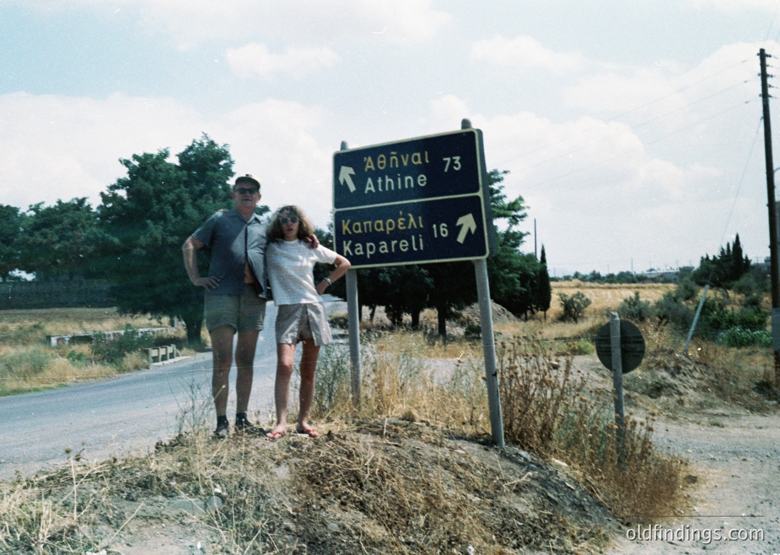 Couple posing beside a Greek road sign marking distances to Athens (73km) and Kaparelli (16km). Mid-20th century attire suggests 1960s-1970s. Rural countryside with dry vegetation and clear skies.