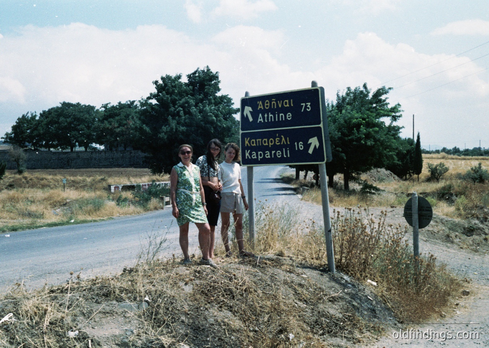 Two women pose beside a Greek road sign (1970s) pointing to Athens (73km) and Kaparelli (16km). Rural countryside with dry vegetation and a winding road. Floral print dress and sunglasses suggest summer attire.