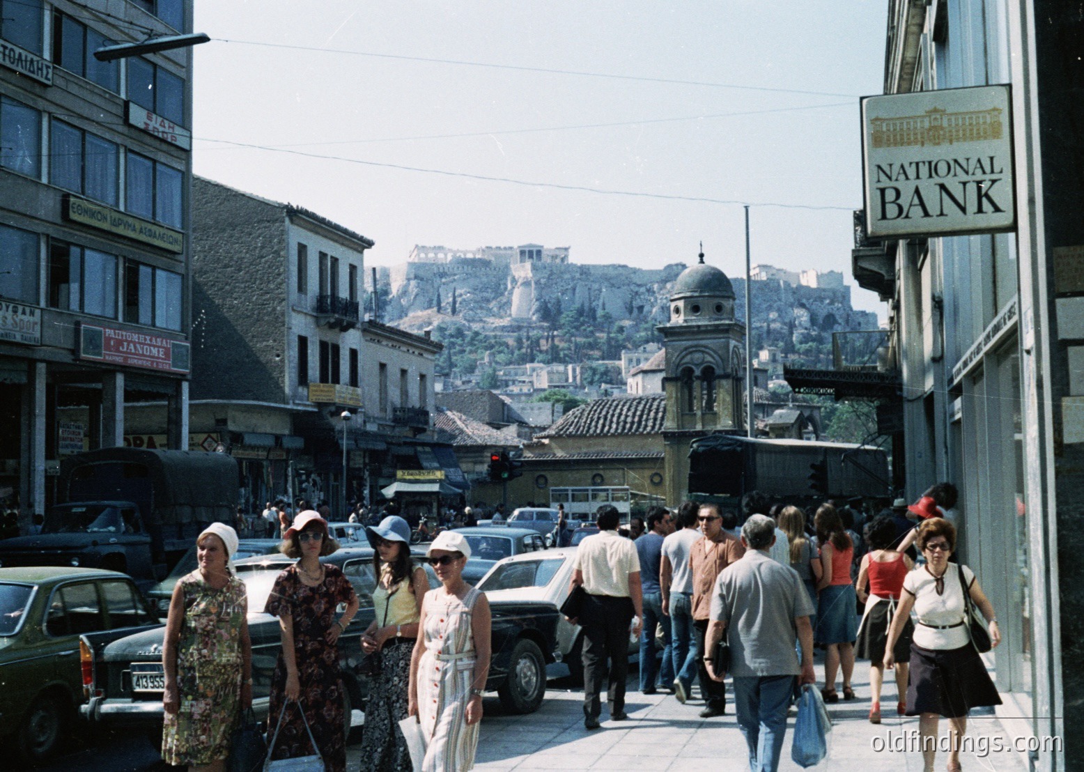 Busy urban street in Athens, Greece, with Acropolis visible in background. Mid-20th century clothing and vintage cars suggest . National Bank sign indicates commercial district. Crowded pedestrian scene with mixed architecture—modernist buildings and traditional elements.
