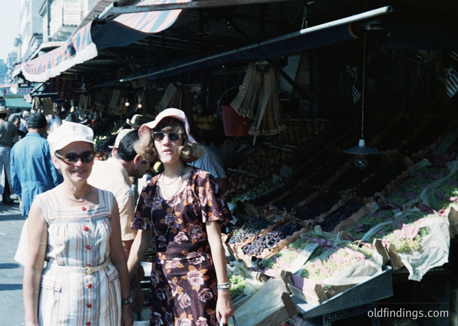 Vintage outdoor market scene featuring two women in 1960s-70s fashion: striped blouse with red buttons and wide-brimmed hat vs. floral dress and sunglasses. Fresh produce and textiles displayed under striped awnings. Urban setting with visible signage in Cyrillic script.