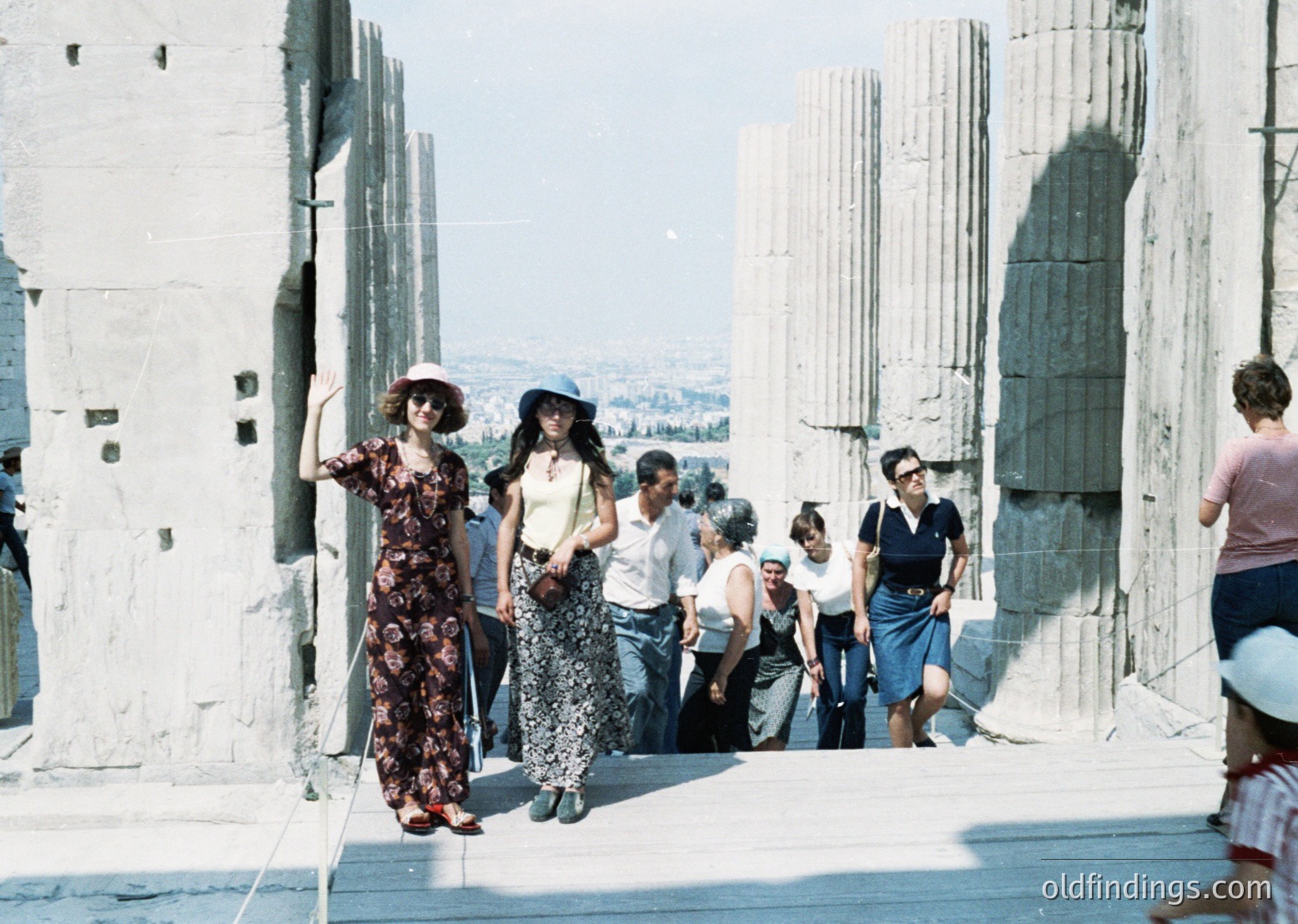 Vintage 1960s photograph of a group walking along the Parthenon ruins in Athens, Greece. Architectural columns and ancient stonework frame the scene. Women in floral dresses, wide-brim hats, and knee-length skirts dominate the foreground, reflecting mid-century fashion. Clear skies and bright lighting enhance the historic site’s grandeur.
