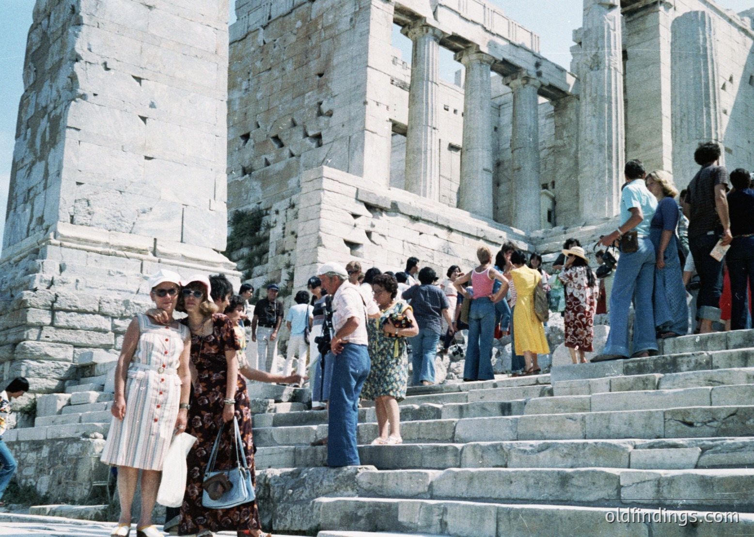 Crowd of tourists ascending ancient stone steps toward the Erechtheion, Acropolis, Athens, Greece, 1960s-70s. Iconic Doric and Ionic columns frame the scene, showcasing classical architecture. Visitors in retro summer attire—flared jeans, floral dresses, and sun hats—highlight mid-century tourism.