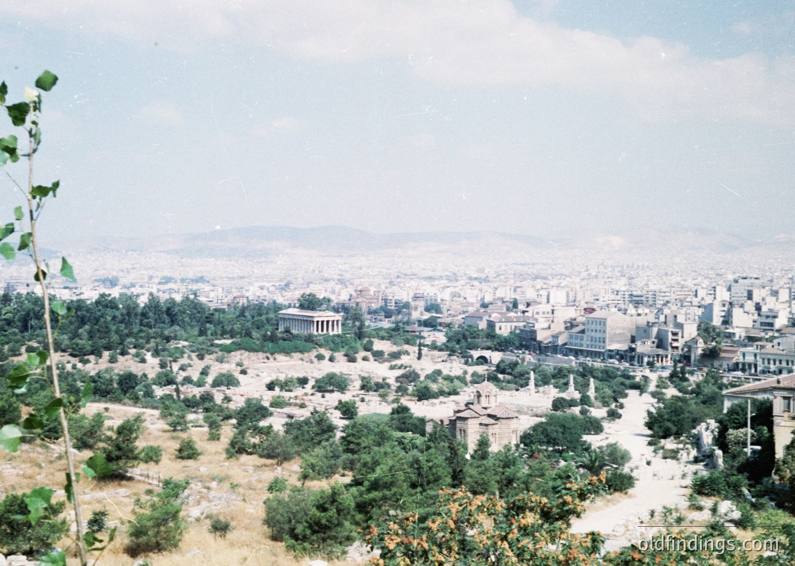 Vintage aerial view of Athens’ ancient ruins blending with modern urban sprawl. The Parthenon and surrounding classical structures dominate the left foreground, framed by sparse greenery. Mid-20th century concrete buildings stretch across the horizon, indicating rapid post-war expansion. Color-tinted film suggests 1950s–1960s era.
