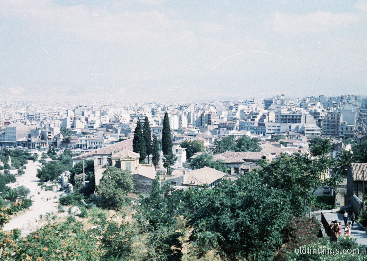Vintage aerial view of Athens’ sprawling urban landscape, blending historic ruins with mid-20th-century architecture. Foreground features lush greenery and a prominent ancient column base, while midground reveals dense residential blocks. Likely captured during the 1950s–1960s, showcasing post-war reconstruction.