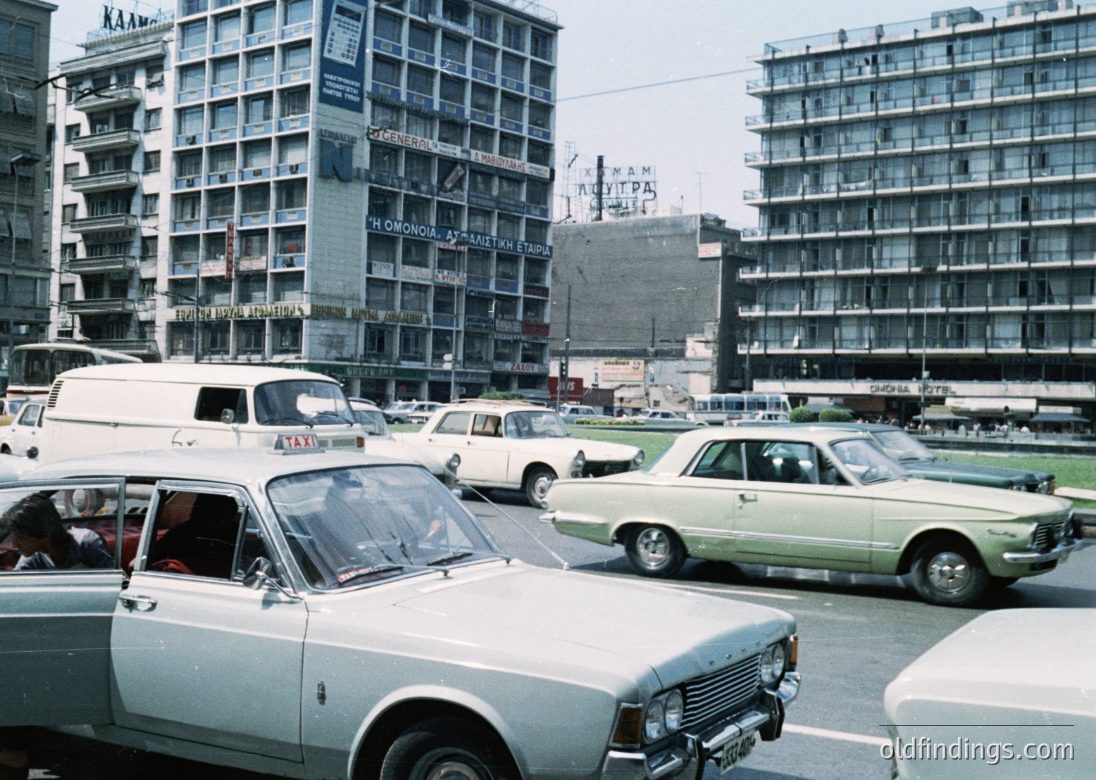 Vintage urban scene featuring mid-rise Soviet-era concrete apartment blocks with Cyrillic signage (). Foreground showcases 1970s-era cars, including a white van and classic sedans (). Busy street with pedestrians, likely a central city plaza (). Colorful but faded, indicative of mid-20th-century photography ().