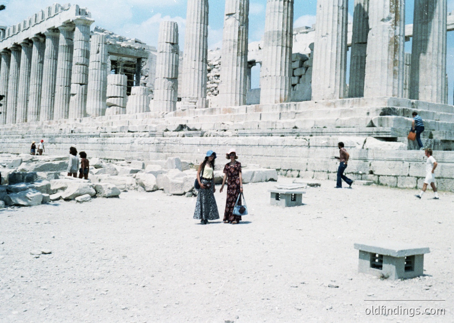 Ancient Greek ruins of the **Temple of Poseidon at Cape Sounion**, Greece, mid-20th century. Weathered marble columns and fragmented architecture dominate the seaside landscape. Two women in traditional attire stand near a concrete bench, while others explore the site.