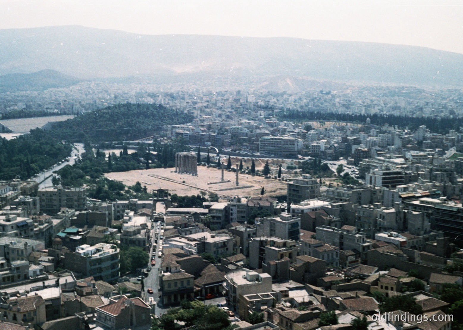Aerial view of the **Ancient Agora of Athens**, showcasing its central ruins surrounded by dense urban sprawl. Mid-20th century architecture blends with classical columns and open plaza. Coastal hills and water body visible in background.