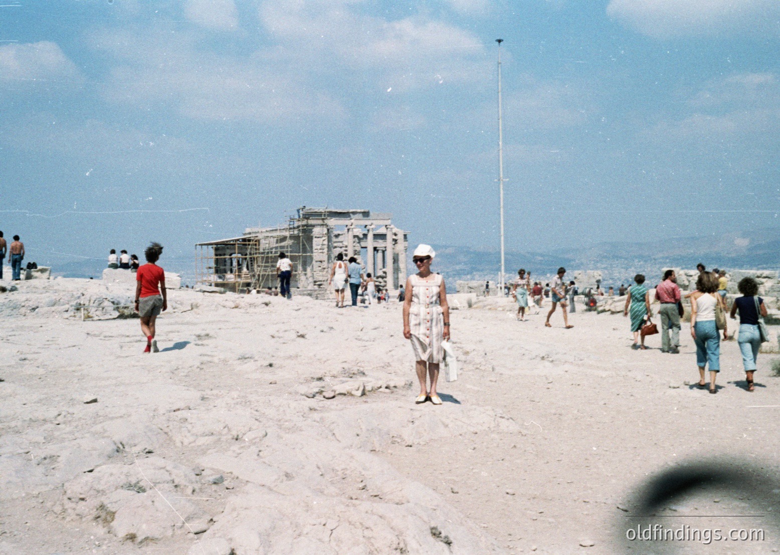 Vintage seaside scene with mid-20th-century beachgoers on rocky terrain. A partially constructed concrete structure (likely a pavilion) dominates the center. Crowds in retro swimwear—shorts, sundresses, and hats—enjoy the sun. Coastal landscape with distant hills under clear skies.