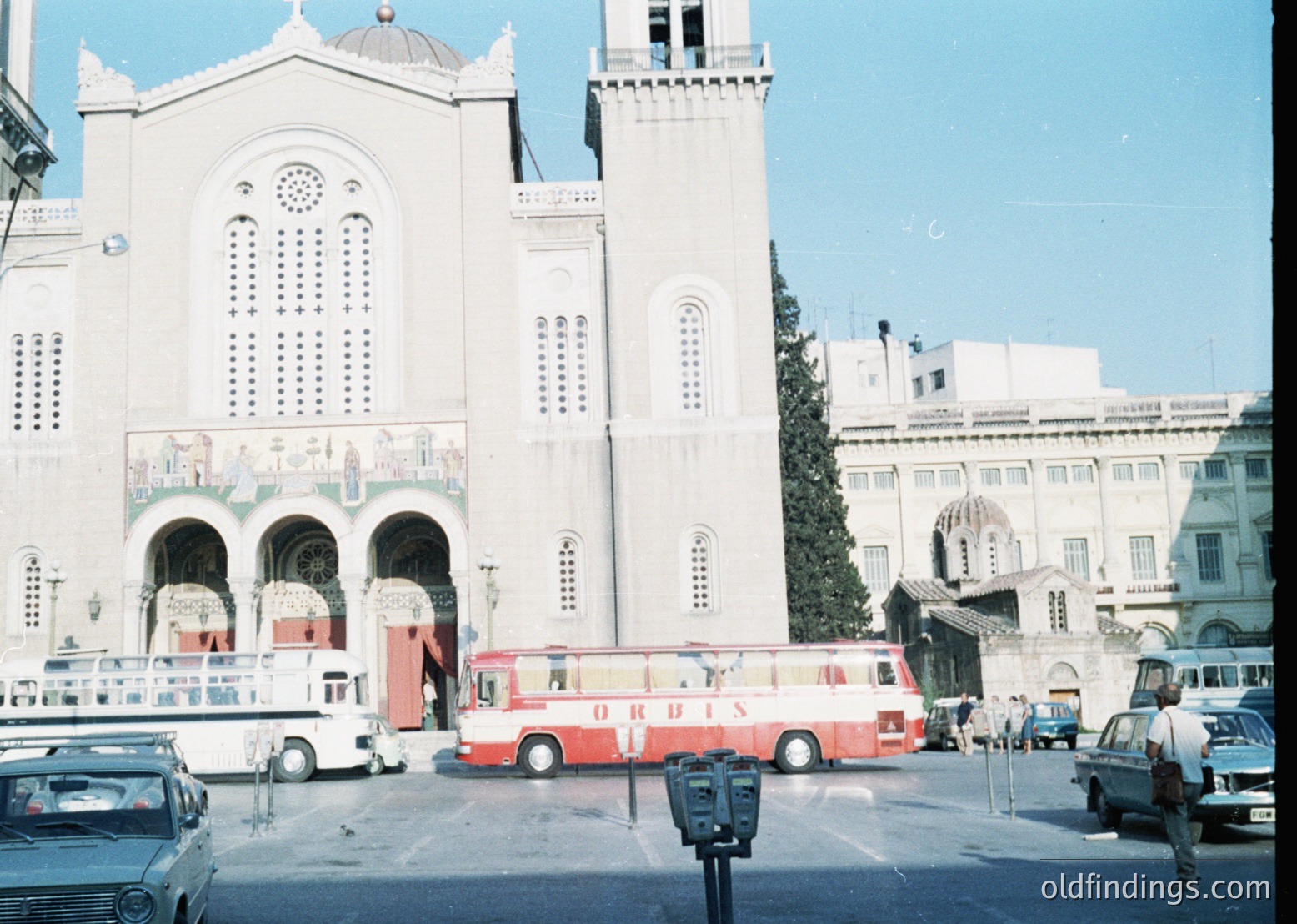 Neoclassical church façade with arched entryways, decorative columns, and circular windows, likely from the mid-20th century. Red "ORBIS" bus and vintage cars in a bustling urban plaza under clear skies. Architectural details include symmetrical design and ornate stonework.