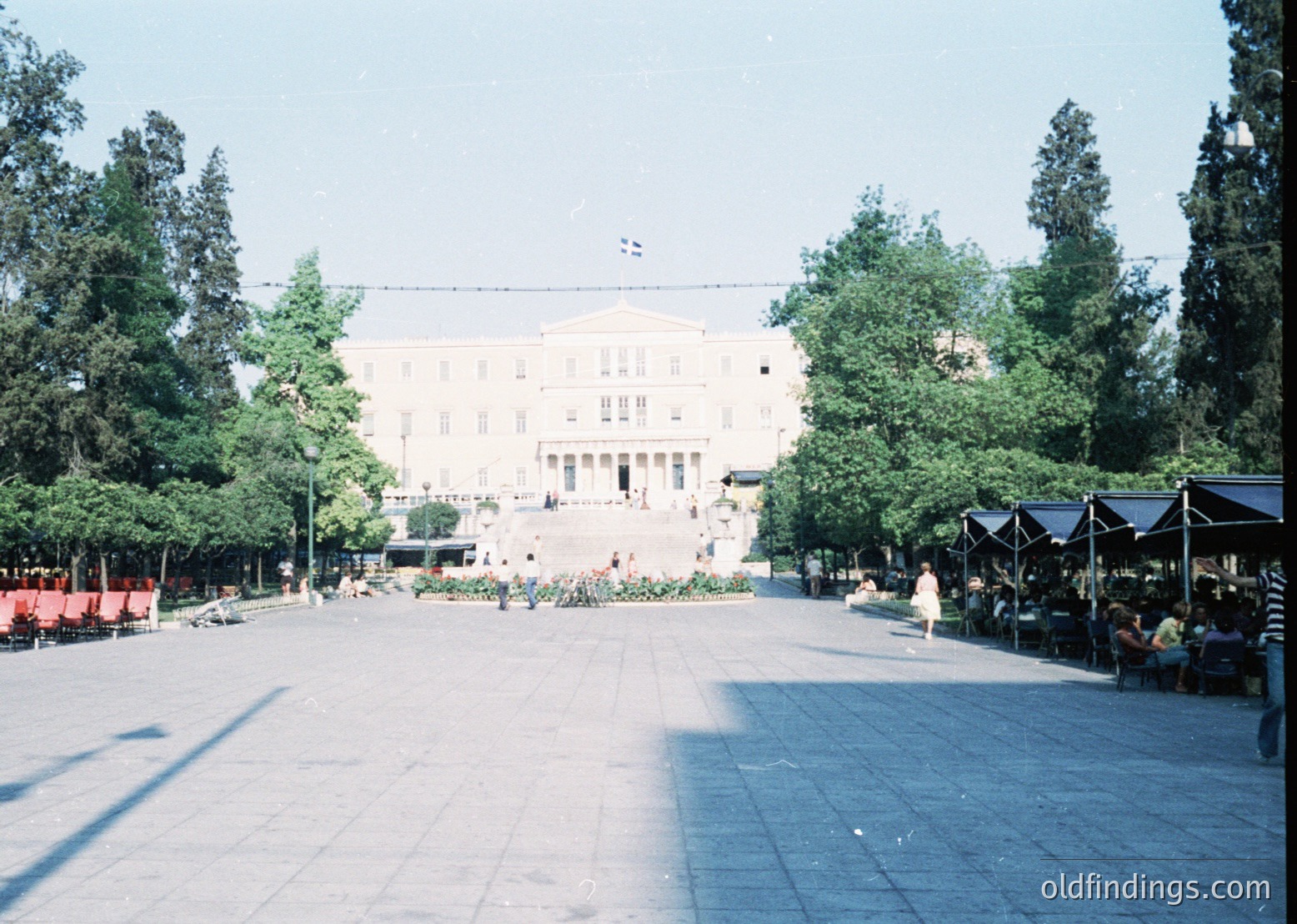Neoclassical government building with symmetrical columns and pediment, flanked by lush greenery and a wide pedestrian plaza. Outdoor market stalls with canopies line the right side. Likely Athens, Greece, mid-20th century.