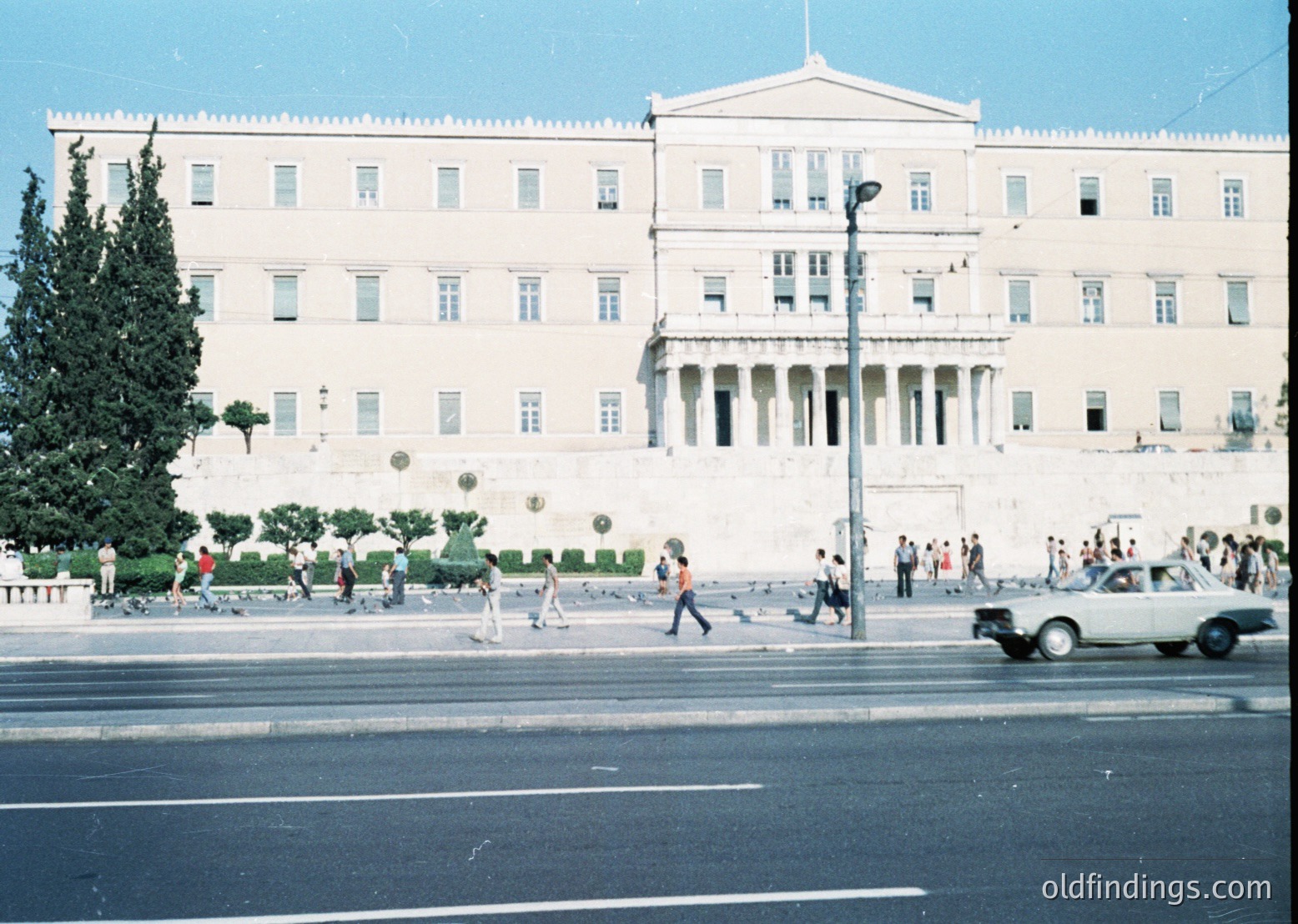 Neoclassical government building with prominent columns and symmetrical façade, likely the Greek Parliament in Athens. Pedestrians and vintage cars populate the wide, tree-lined street ( )