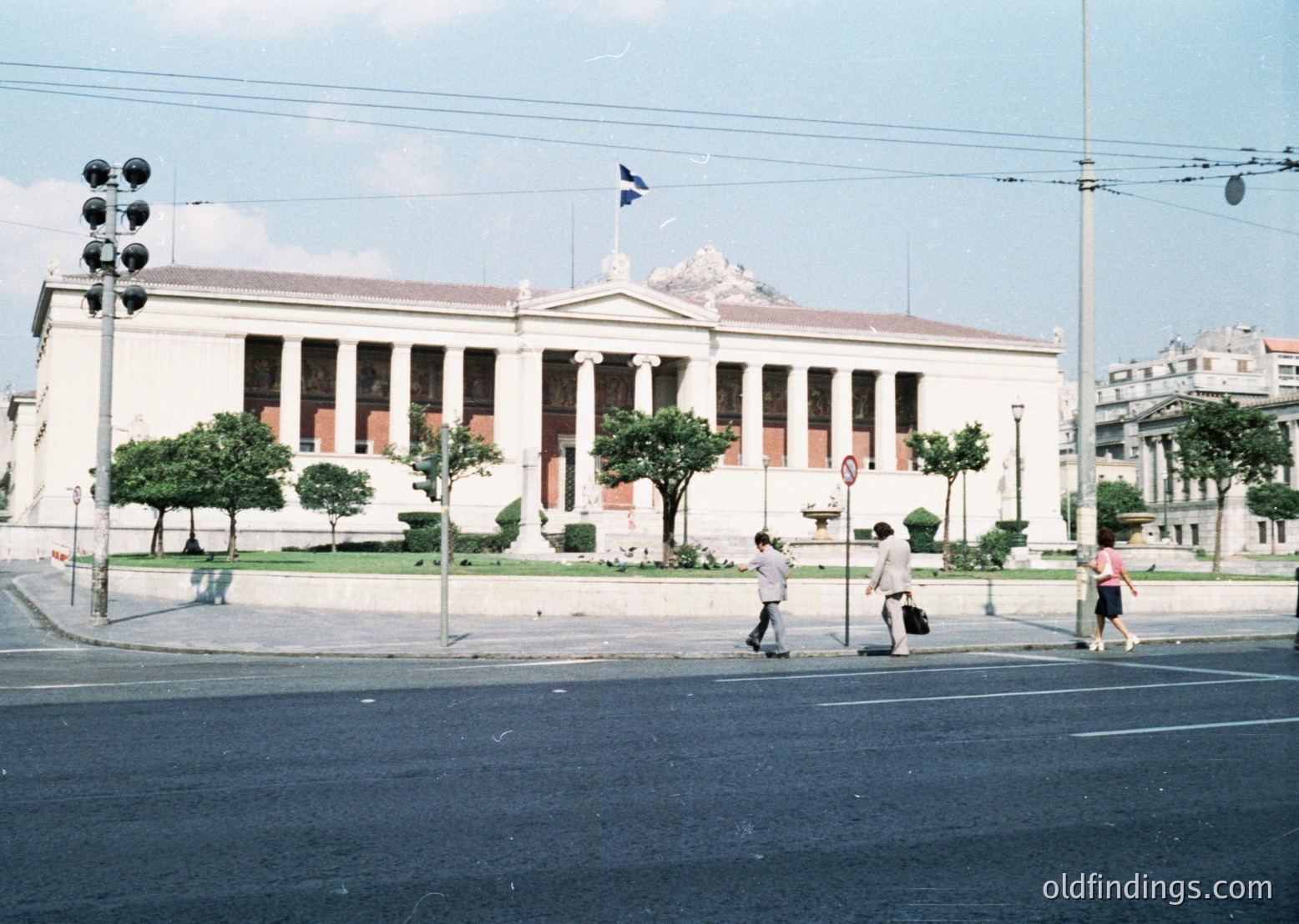 Neoclassical building with prominent columns and pediment, likely a government or cultural institution, set against a clear sky. Greek flag atop, suggesting Athens, Greece. Pedestrians crossing street in mid-20th century attire (). Urban landscape with streetlights, trees, and distant mountains.
