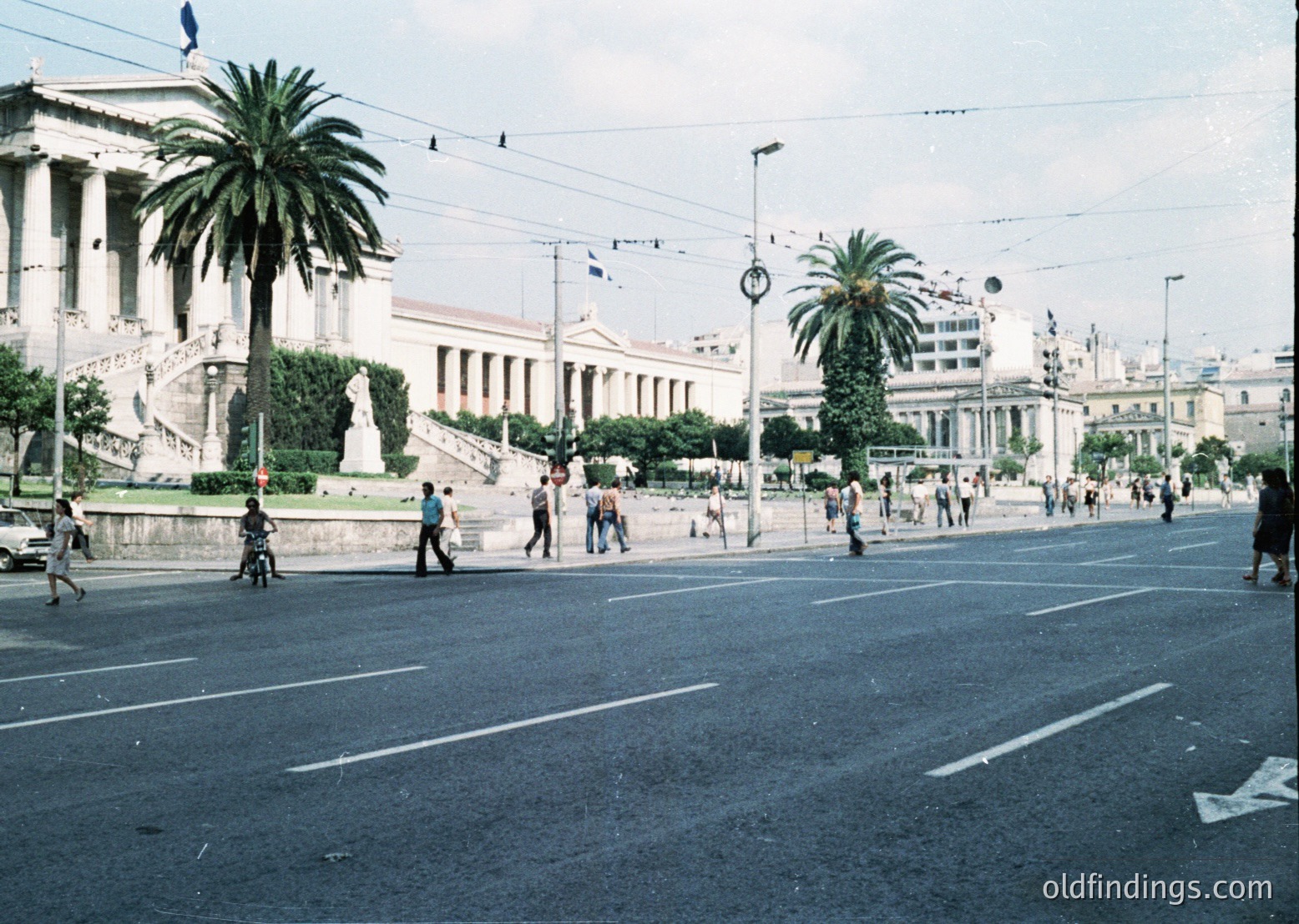 Neoclassical building with grand columns and steps in urban setting, flanked by palm trees. Mid-20th century street scene with pedestrians, motorcycles, and overhead wires. Likely Athens, Greece (-1960s).