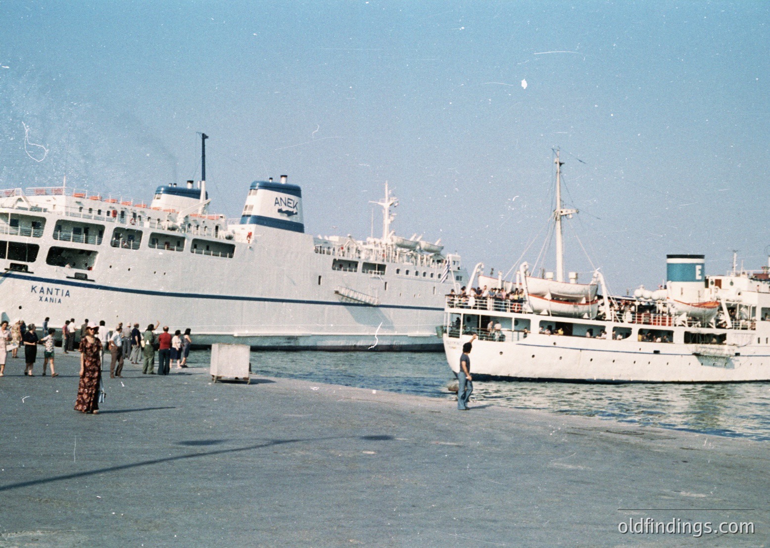 Mid-20th century ferry port scene: Two large passenger ships ("Kantia" and "ANEX") docked at a bustling pier with crowds boarding/disembarking. Bright, sunny weather with clear skies.