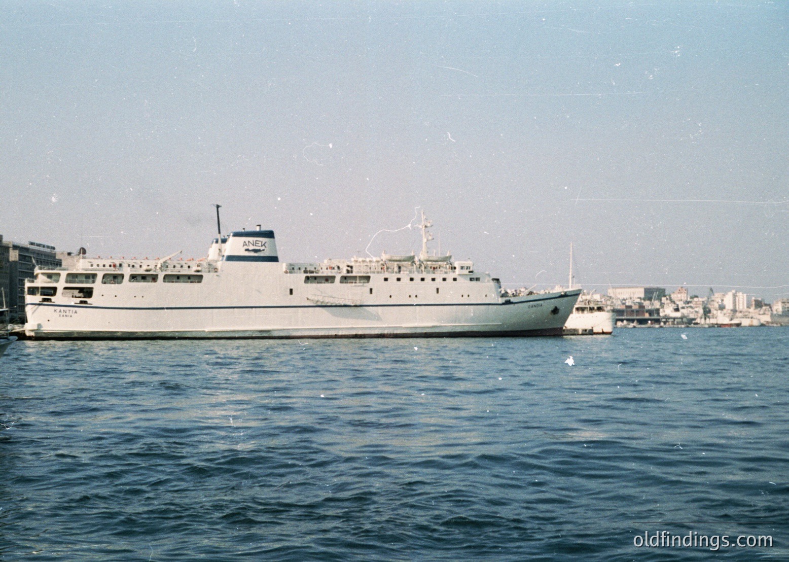 Mid-20th century ferry docked at port, likely Mediterranean. White hull with "Aegean" lettering, multiple decks, and lifeboats visible. Urban coastline with low-rise buildings in background. Vintage grain and color tone suggest 1960s–1970s era.