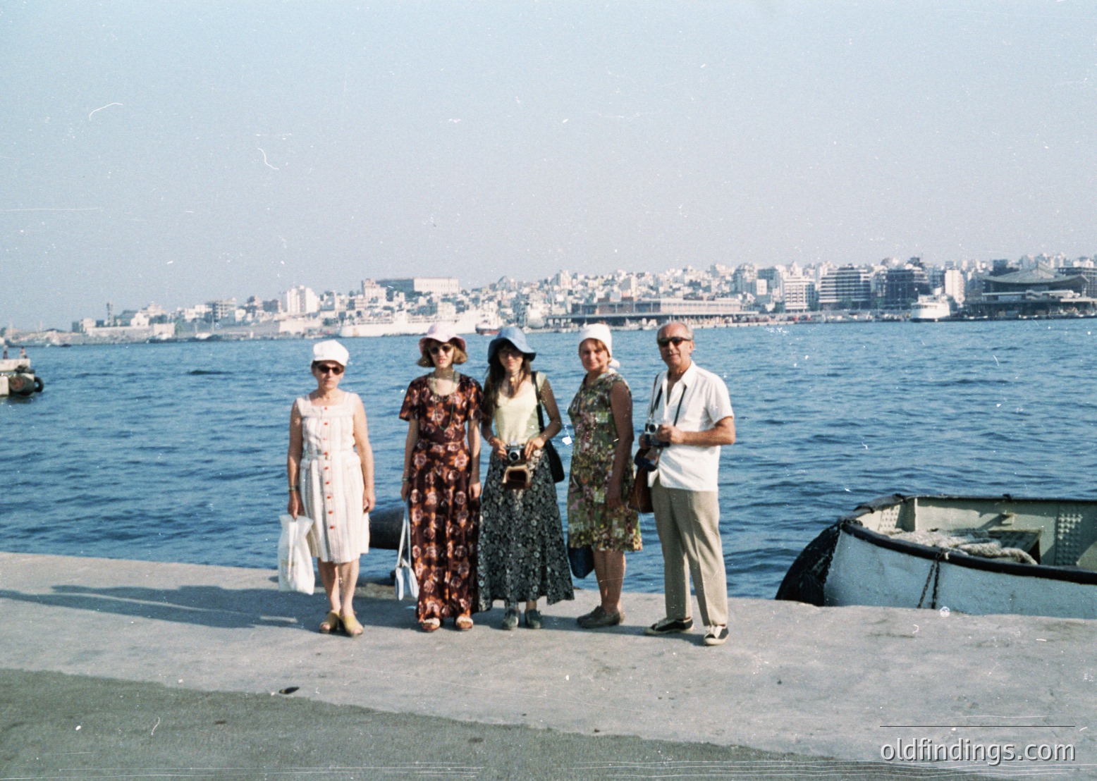 Five individuals pose on a seaside promenade, likely Istanbul’s Golden Horn, 1960s–1970s. Women wear mid-century dresses with floral/patterned fabrics; one holds a camera. Urban skyline and waterfront boats frame the scene.