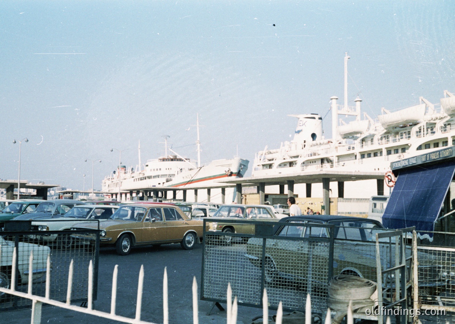 Vintage ferry port scene with mid-20th century passenger ships docked. Yellow classic car (likely a 1970s Ford Escort) parked in a fenced lot beside older vehicles. Open-air terminal with covered walkways and maritime infrastructure. Clear skies, suggesting coastal or seaside location.