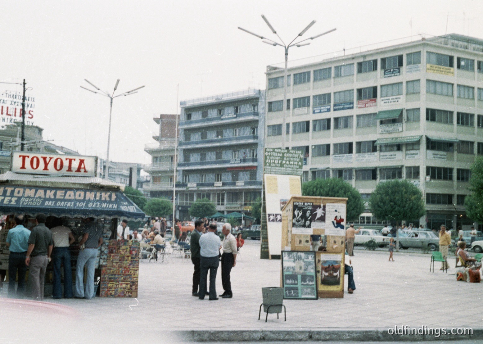 Vintage urban street scene featuring mid-20th century Thessaloniki, Greece. A Toyota dealership sign and "Tom Makedoniki" (automotive parts) stall dominate left. Pedestrians in 1970s-era clothing walk past a newsstand displaying magazines and posters. Concrete high-rise buildings with balconies and signage in Greek frame the scene. Classic cars and vintage streetlamps add to the era’s authenticity.