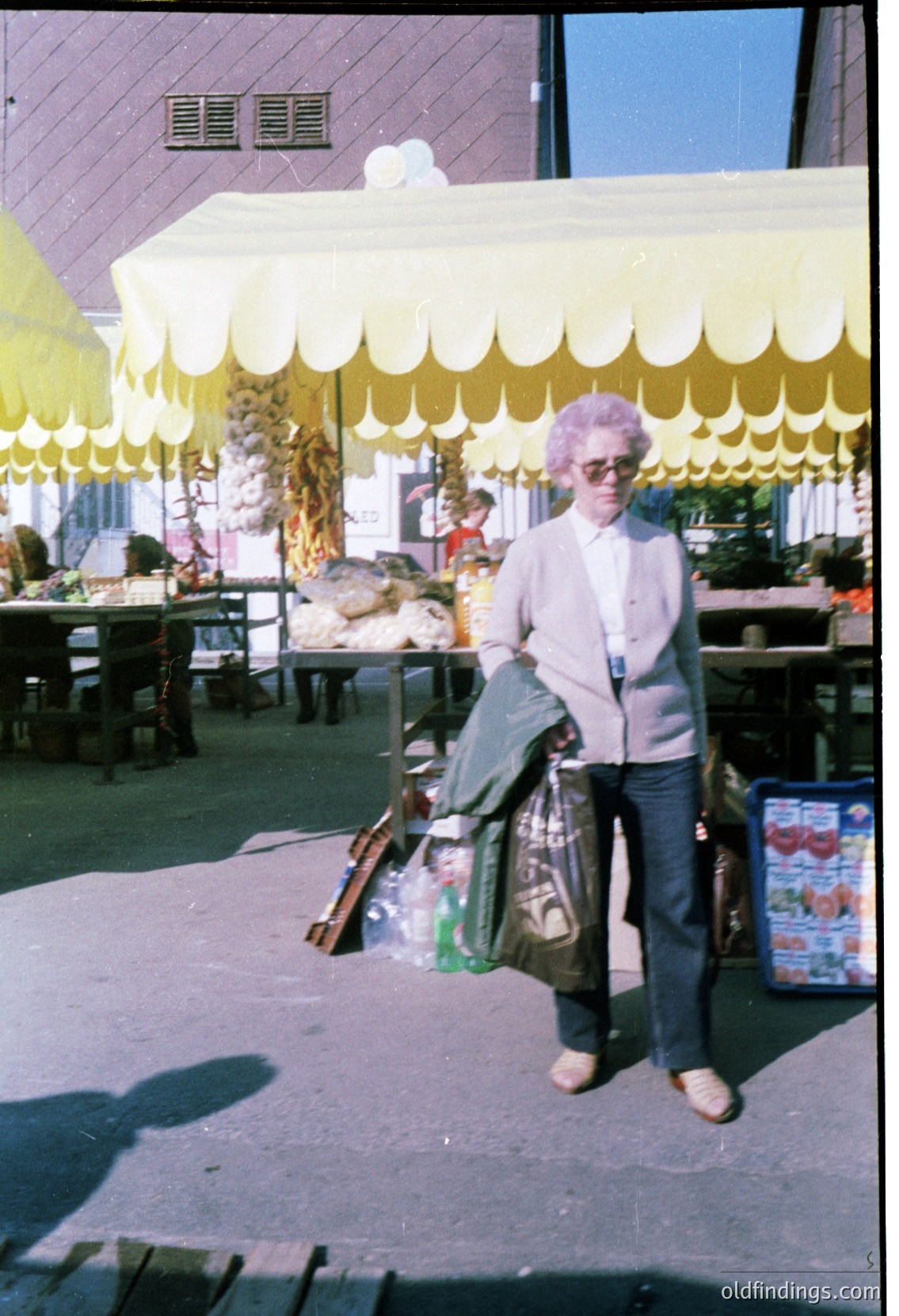 Vintage outdoor market scene featuring an elderly woman in a purple wig, sweater, and apron carrying a bag and broom. Yellow scalloped canopy tents and wooden tables with goods (including a visible "Smetana" container) suggest a Soviet-era or Eastern Bloc market setting, likely 1960s–1980s.