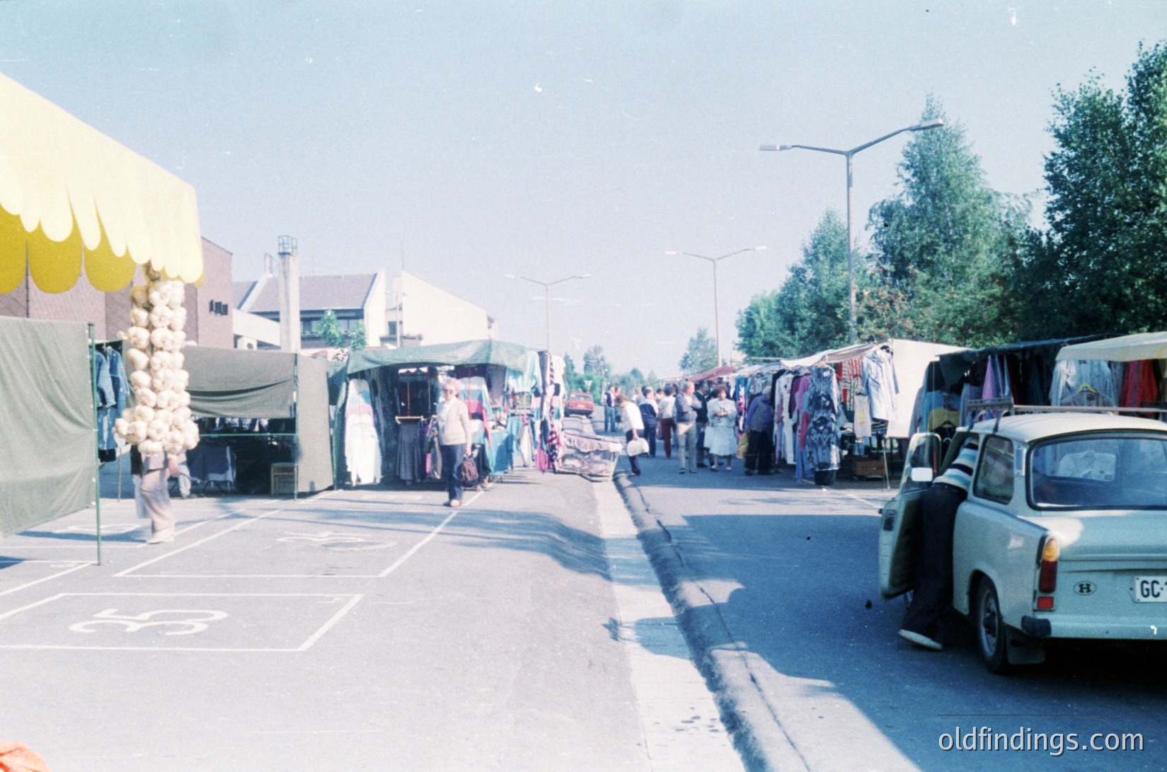 Vintage outdoor flea market with makeshift stalls displaying vintage clothing, household items, and textiles. Narrow road flanked by parked cars, including a classic white sedan. Brightly colored canopies and draped fabrics create a bustling, nostalgic atmosphere. Likely Eastern European setting, mid-20th century.