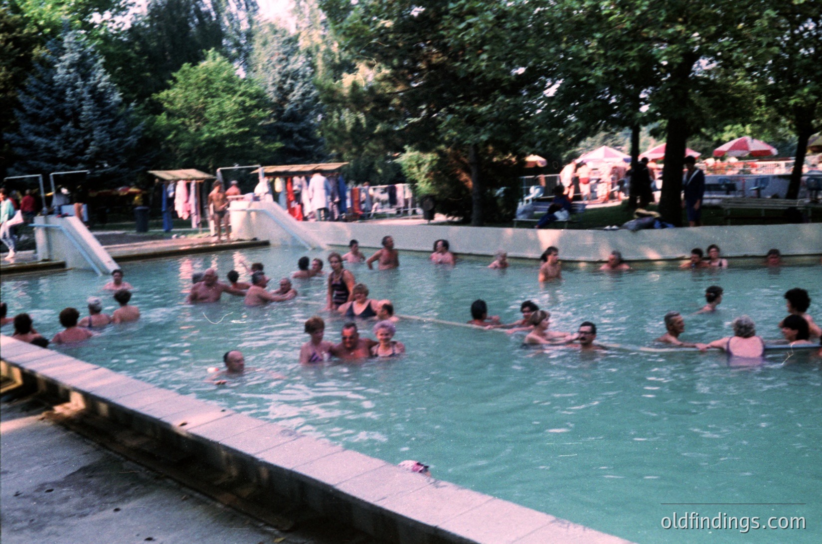 Vintage outdoor swimming pool filled with people in swimsuits, likely mid-20th century. Lush greenery surrounds a shallow, rectangular pool with a water slide. Crowded, lively atmosphere suggests a public or resort setting.