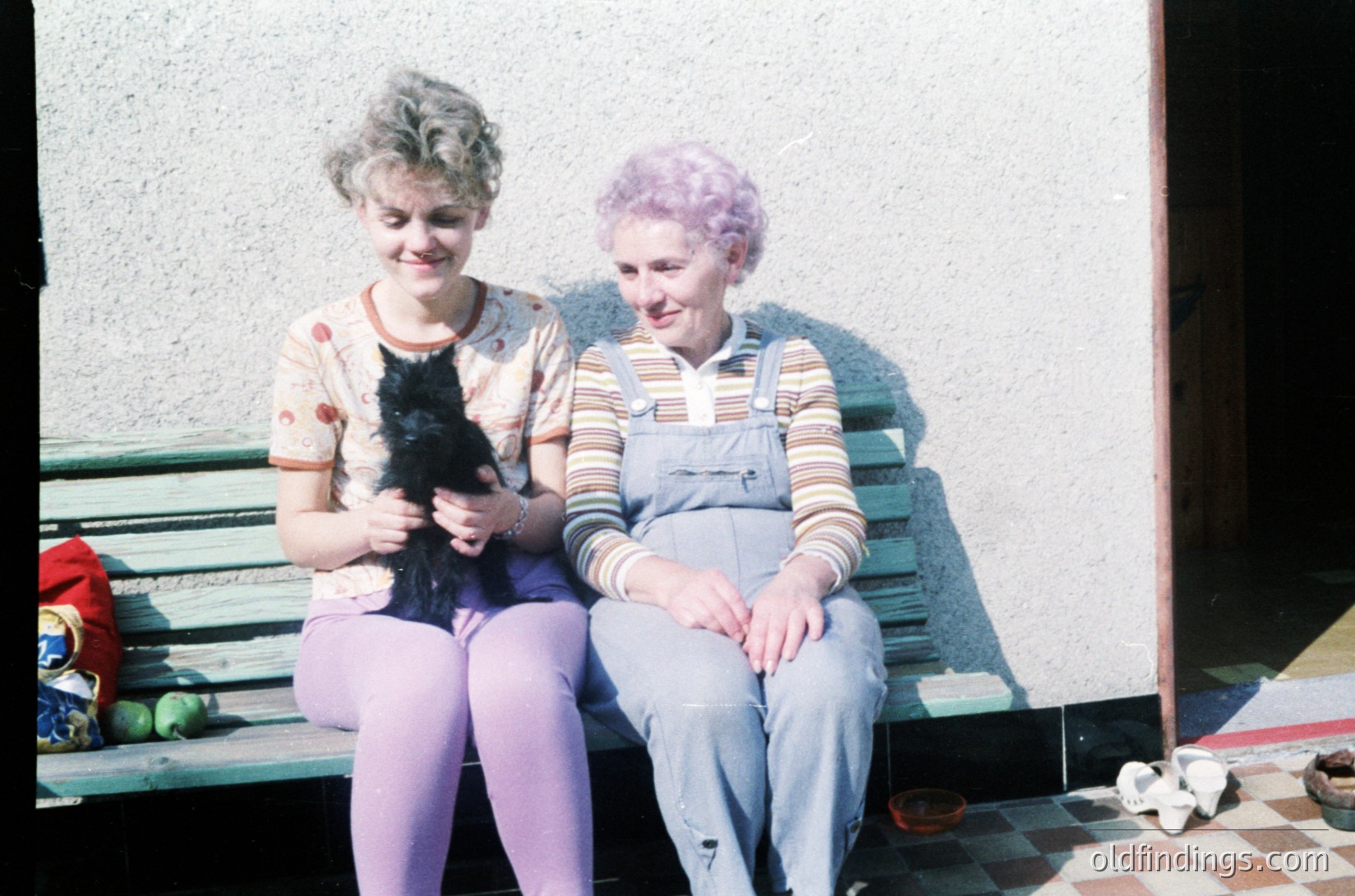 Two women sit on a wooden bench holding a black cat, likely from the 1970s. The woman on the left wears a floral blouse, purple tights, and a headband; the right wears striped overalls and a headscarf. The setting appears domestic, with a tiled floor and a doorway in the background.