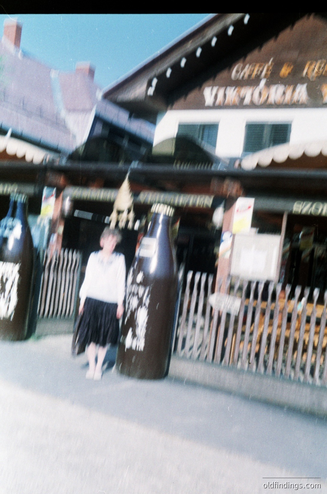 Vintage photo of a child in a white blouse and dark skirt posing beside large glass beer bottles at a seaside café, "Café Victoria." The setting appears to be a European coastal town, likely the 1960s–70s. The blurred background suggests motion or low-resolution film. é