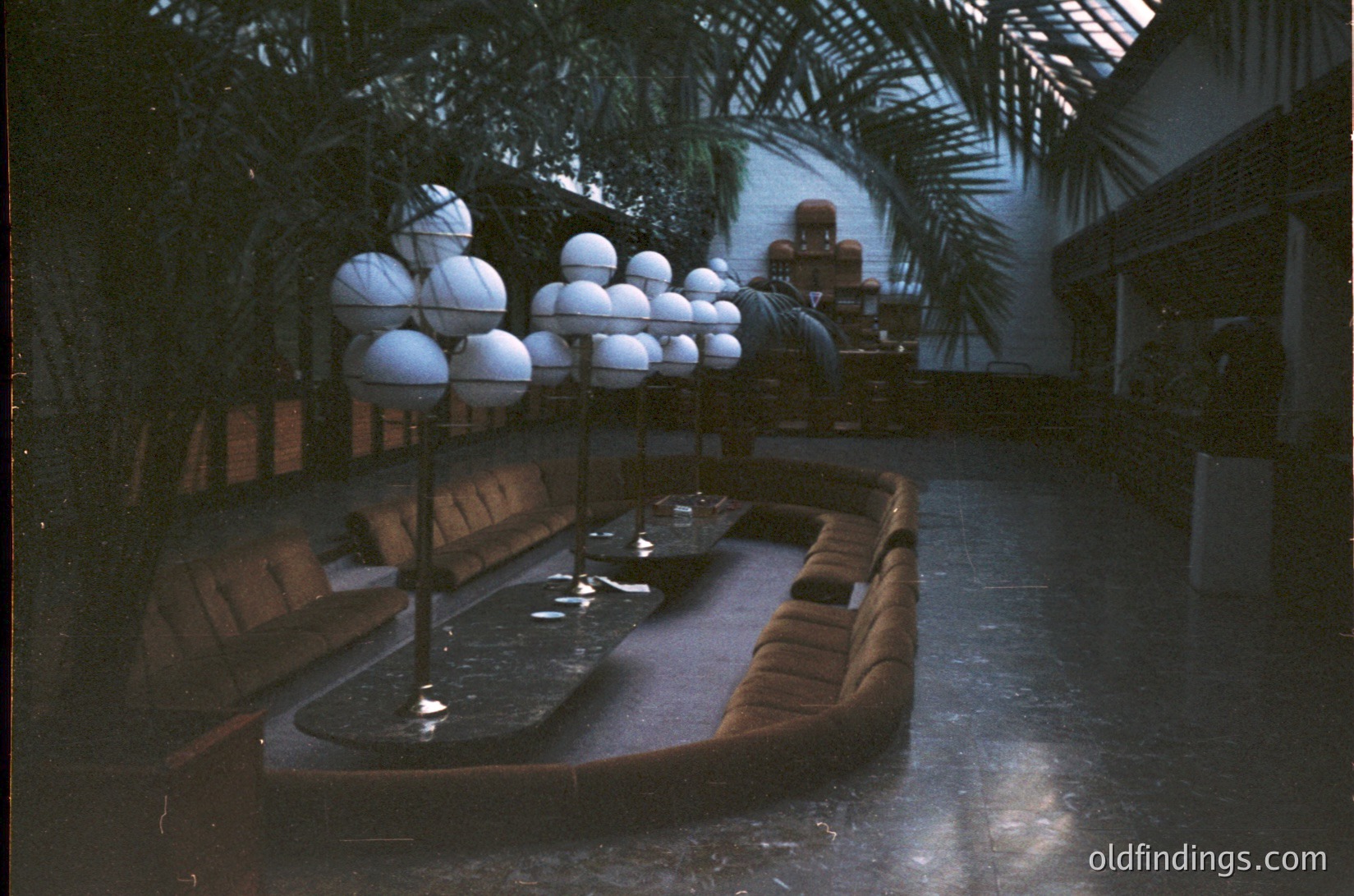 Mid-century modern indoor courtyard with curved leather seating, central shallow pool, and spherical light fixtures. Lush tropical plants frame the space, blending natural and architectural elements. Likely a 1960s-70s hotel or resort lobby.