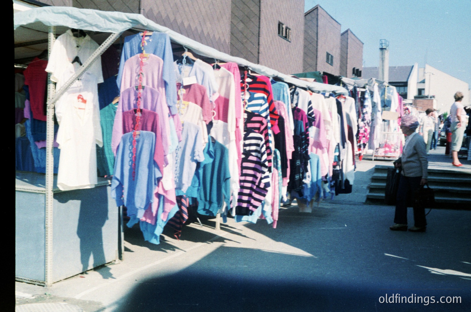 Vintage outdoor clothing market stall with colorful 1970s-style garments—striped, floral, and solid prints—displayed on lines. Urban setting with concrete barriers and a modernist building in background. A man in dark pants examines items. Retro fashion research or nostalgic design reference.