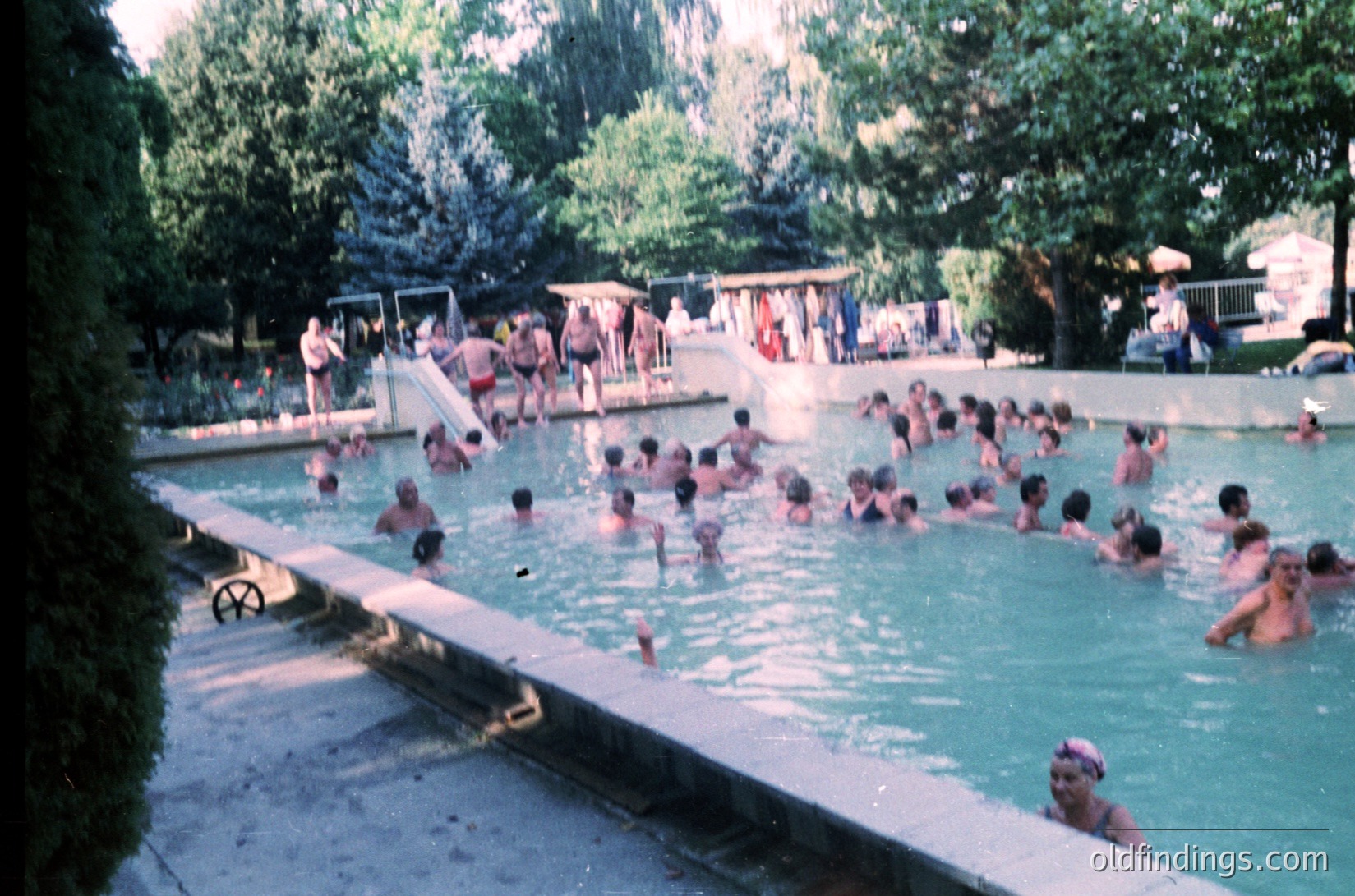 Vintage outdoor swimming pool filled with people, likely from the 1960s–1970s. Concrete edges, shallow diving platform, and lounge chairs under leafy trees. Casual summer attire and relaxed social atmosphere.