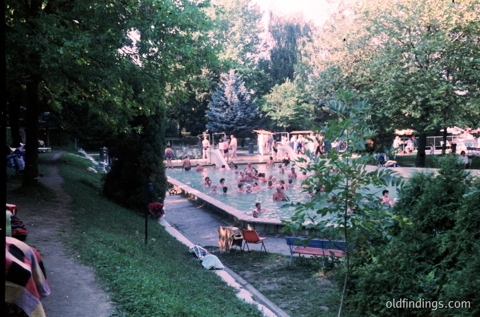 Vintage outdoor swimming pool surrounded by lush greenery, likely from the 1970s. Concrete steps lead into shallow and deep sections, with wooden decking and changing cabins in background. Sunbathing and swimming activities visible.