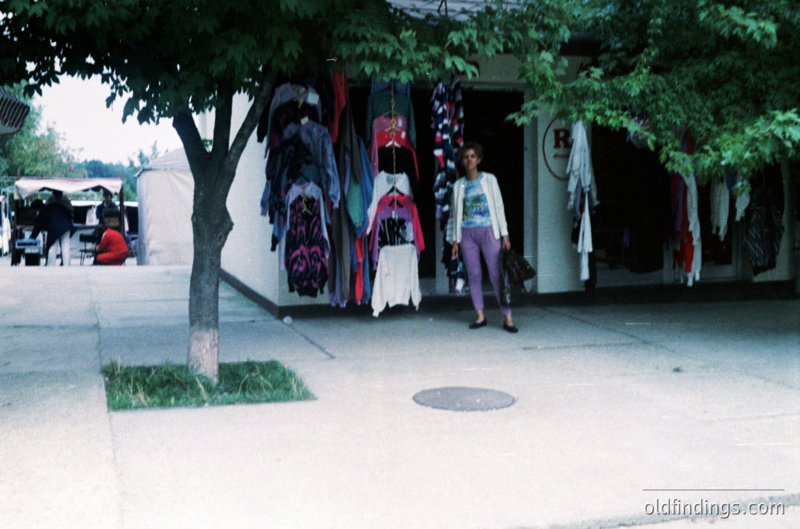 Vintage outdoor clothing stall with colorful, patterned garments hung on display. Woman in 1970s-style patterned top and wide-legged pants walks past. Tree and manicured sidewalk with manhole cover in foreground. Likely a flea market or street fair setting.
