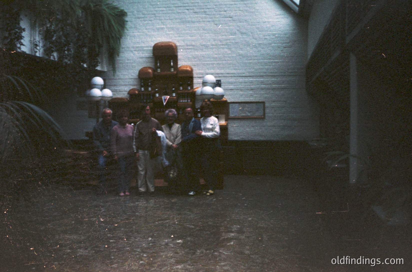 Vintage photo of a group gathered around a vintage jukebox in an indoor courtyard. The setting appears dimly lit with a brick wall and potted plants. Likely 1960s–1970s era, suggesting a social or recreational space.