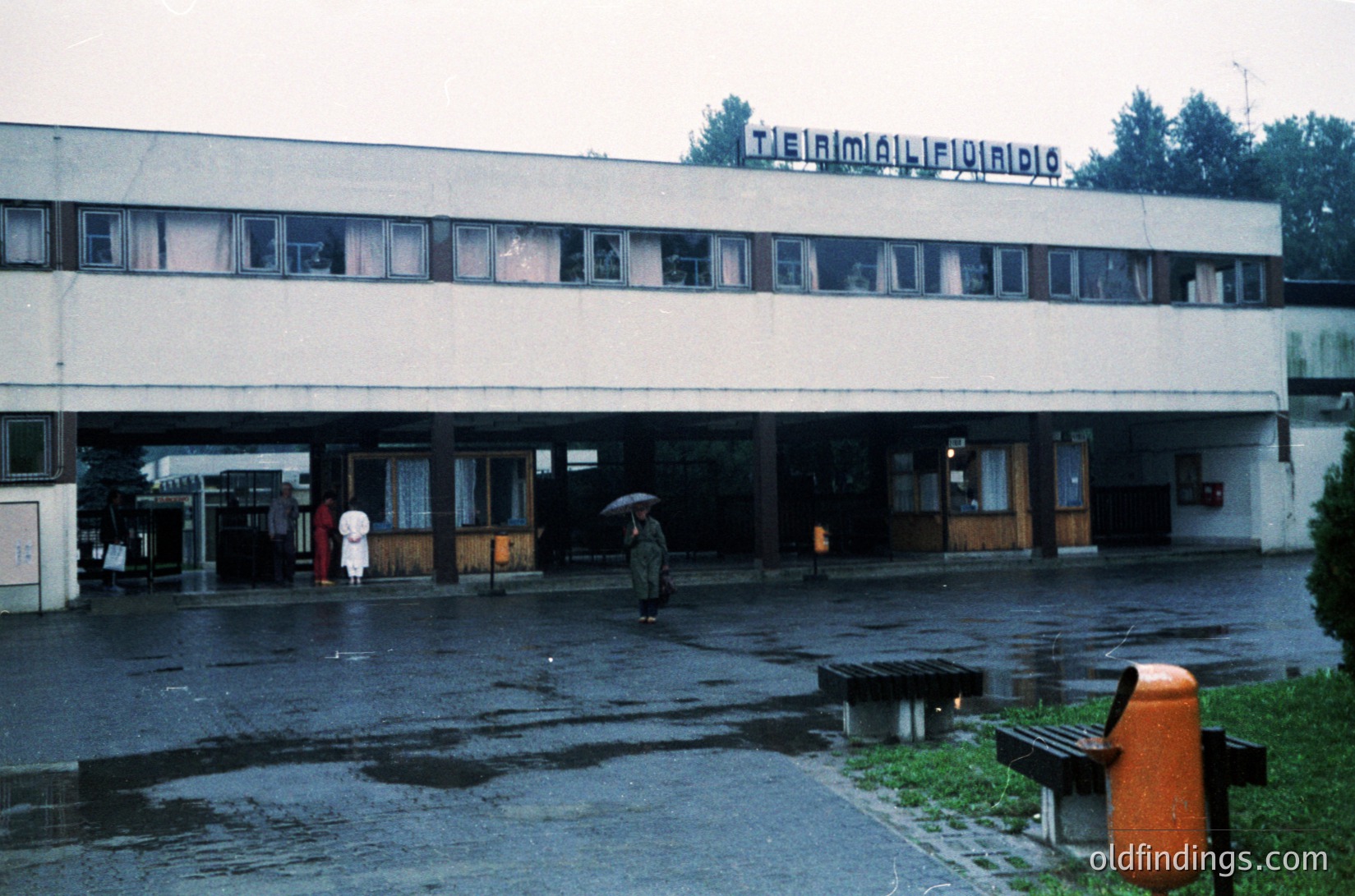 Mid-century institutional building with brutalist concrete elements, featuring a sign reading *"ТЕХНИКУМ МЕХАНИЗАЦИЯ СЕЛСКОТО СТОЧАРСТВО"* (Bulgarian for Agricultural Mechanization Technical School). Wet pavement reflects muted light; two pedestrians—one holding an umbrella—enter/exit via open garage-style entrance. Orange trash bin and concrete benches in courtyard. Likely Bulgaria, 1960s–1980s.