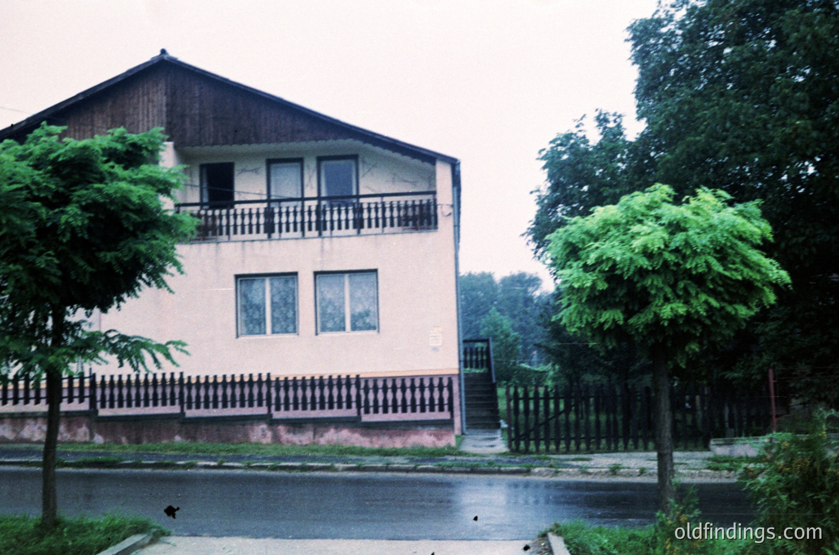 Two-story residential building with mid-century modern elements: cream-painted walls, dark wooden balcony railings, and a sloped roof. Decorative black-and-white striped trim under windows. Surrounded by mature trees and a gated driveway. Likely Eastern European suburban architecture, 1960s-1970s.