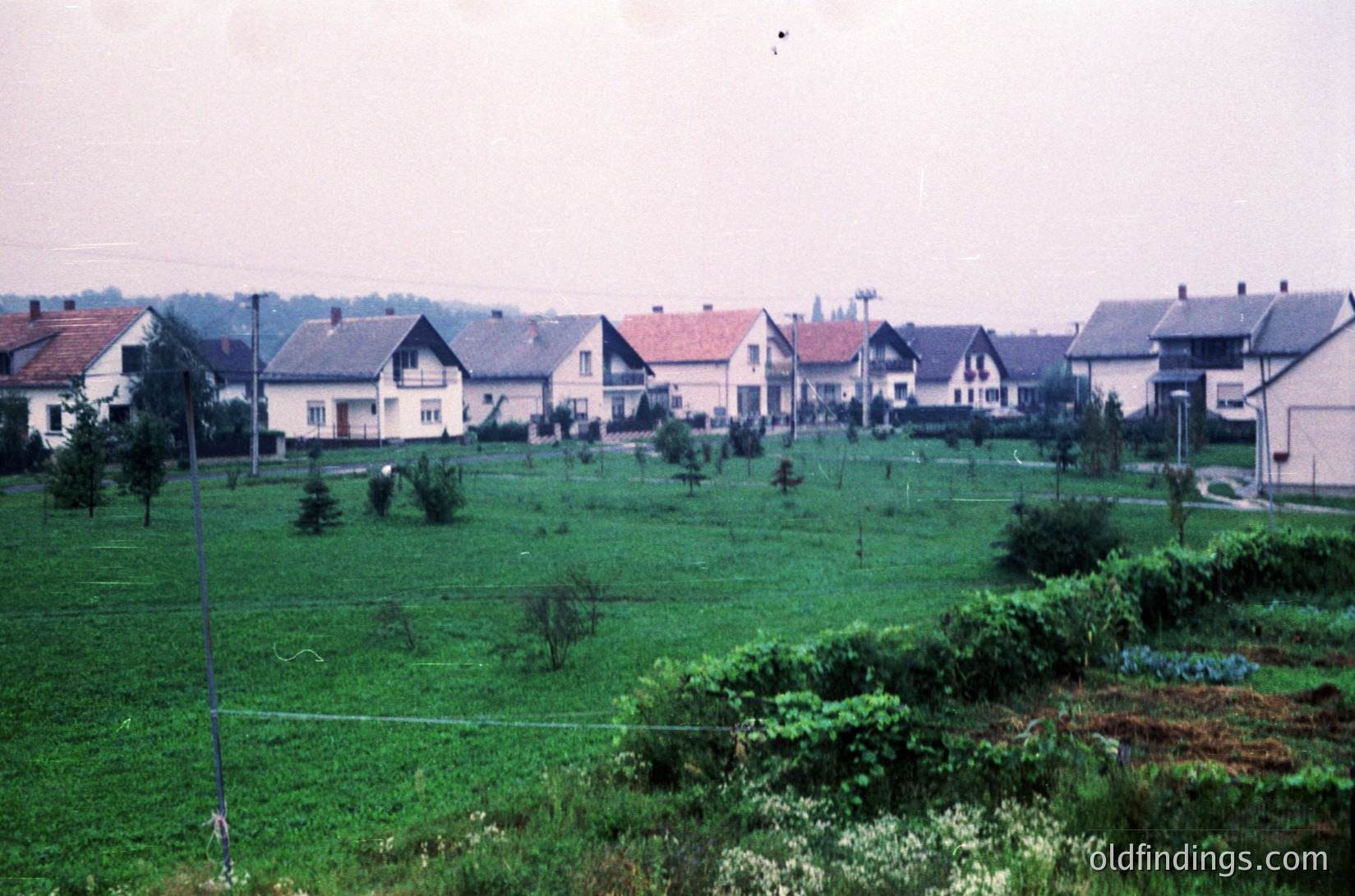 Mid-20th century suburban housing row with uniform brick and stucco facades, gabled roofs, and small front yards. Lush green lawns and scattered trees in foreground suggest communal or private gardens. Overcast sky enhances muted tones. Likely Eastern European post-war housing development.