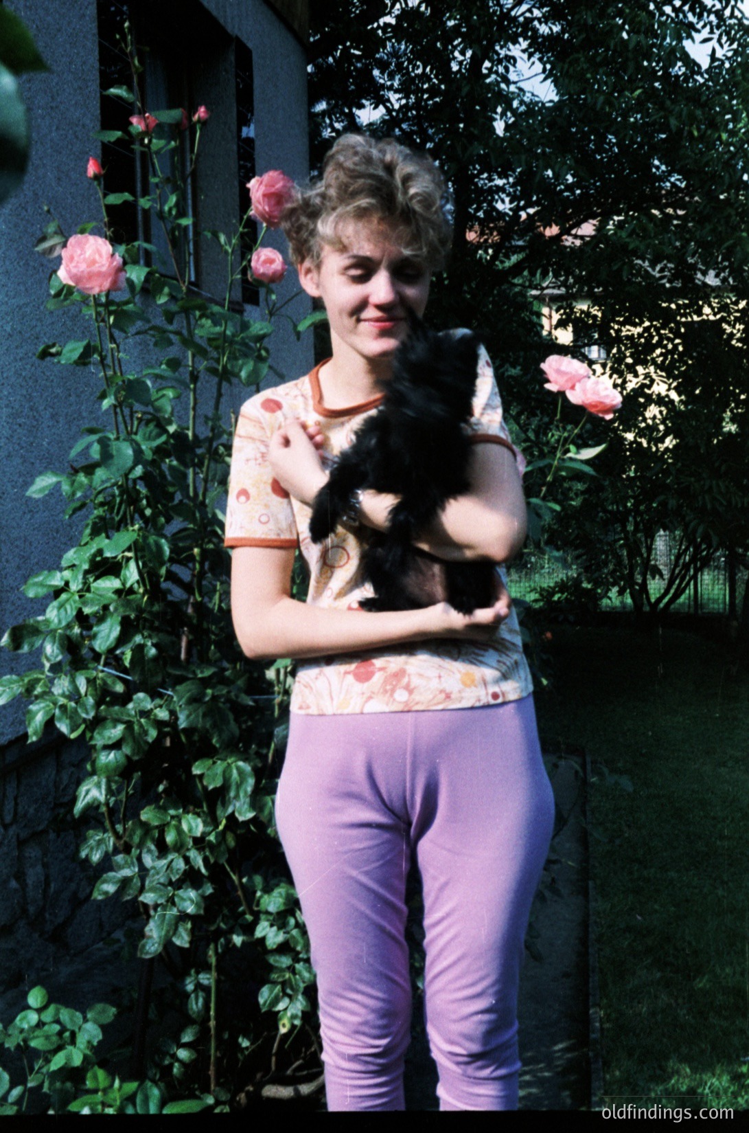 Vintage portrait of a person holding a black cat amidst lush pink roses, likely 1970s–1980s. Floral-patterned top and purple pants suggest retro fashion. Soft focus and warm tones evoke nostalgic family photography.