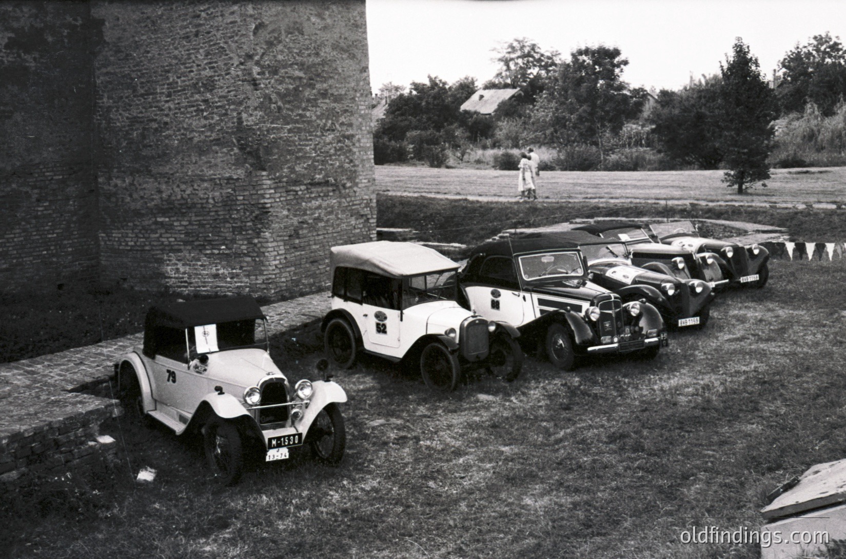 Vintage lineup of classic cars parked beside a stone wall, likely mid-20th century. Open-top models dominate, featuring rounded fenders and minimalist designs. Rural setting with sparse vegetation and distant figures.