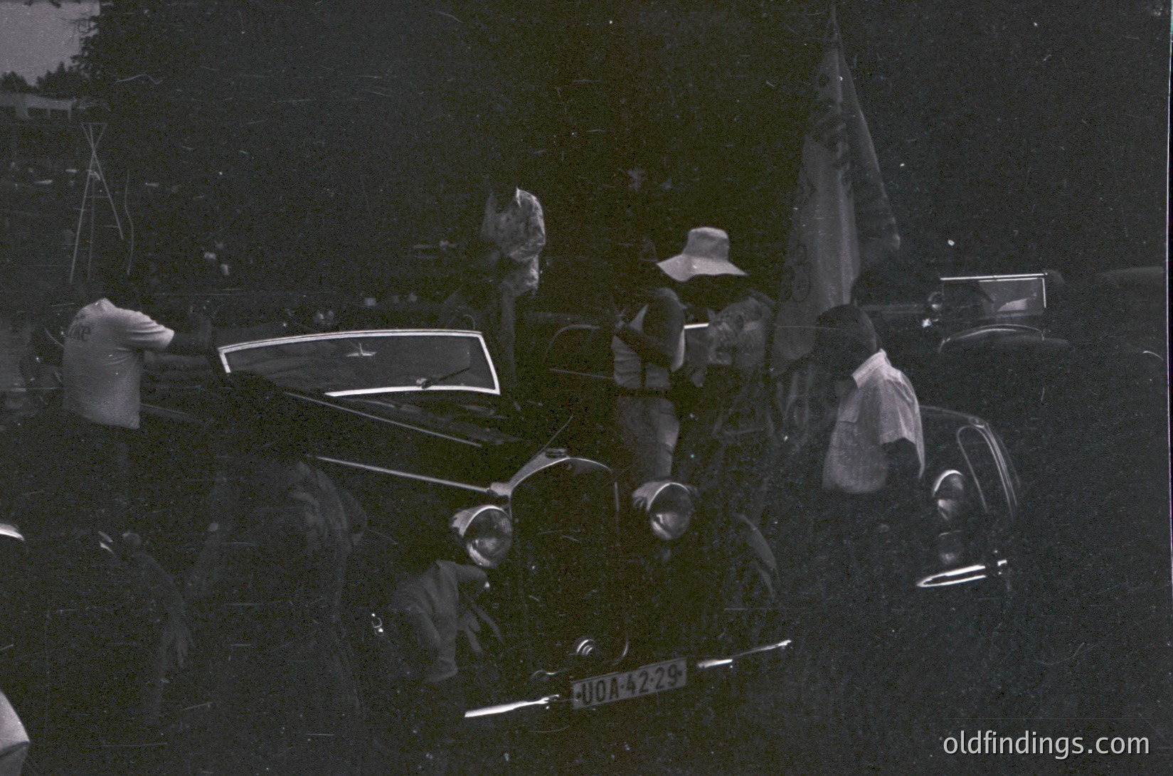 Black-and-white street scene featuring vintage car (license plate "J044229") with passengers in 1940s-50s attire. Crowd gathers around, some holding flags or banners. Urban setting with visible buildings and parked vehicles. Evokes mid-century European public celebration or parade.