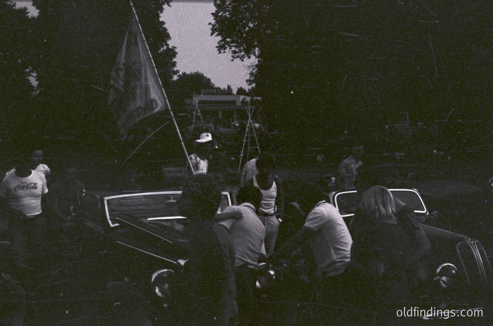 Vintage black-and-white photo of a mid-20th-century outdoor gathering. Group of men in casual 1950s-60s attire (white shirts, dark pants) pose around a vintage car with a Coca-Cola logo on the driver’s shirt. A flagpole with an indistinct emblem stands behind them, suggesting a community or event setting. Trees and a trailer in the background hint at a rural or suburban picnic area.