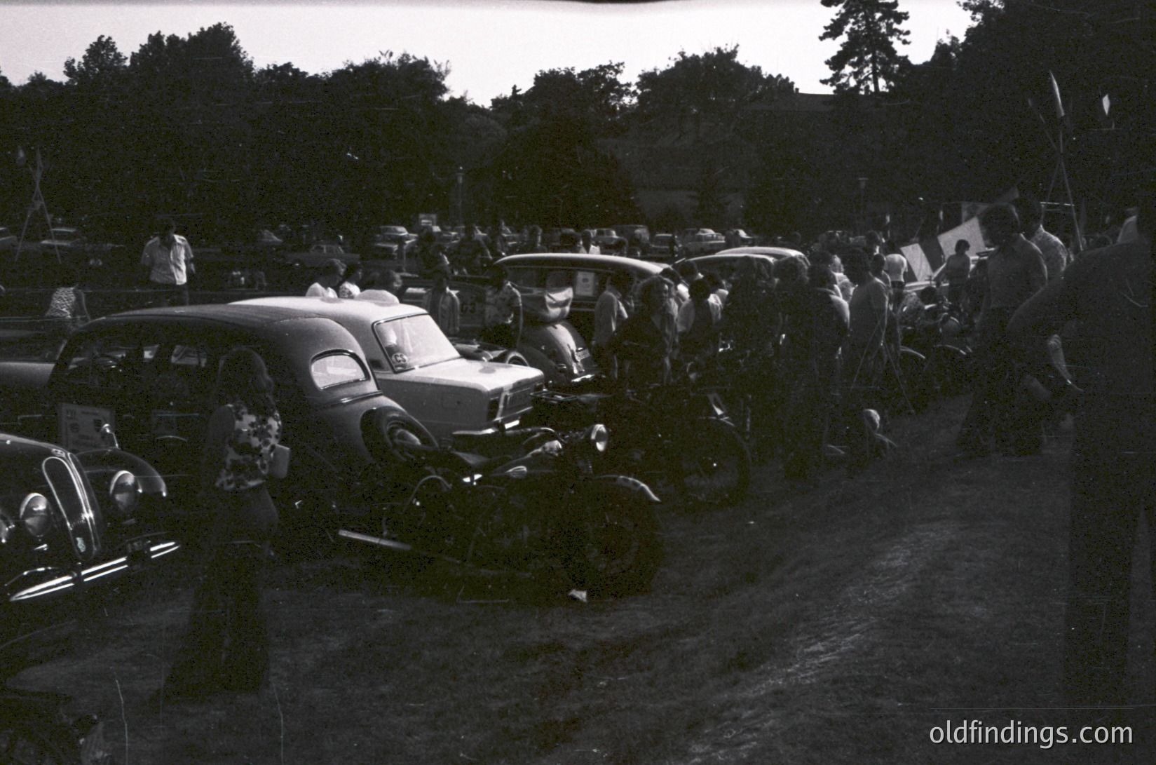 Vintage black-and-white gathering of classic cars and motorcycles in a rural outdoor setting, likely mid-20th century. Packed lineup of pre-1960s vehicles, including streamlined sedans and vintage motorcycles, parked closely. Trees and greenery frame the scene, suggesting a countryside or small-town event. Crowd of onlookers visible in background.