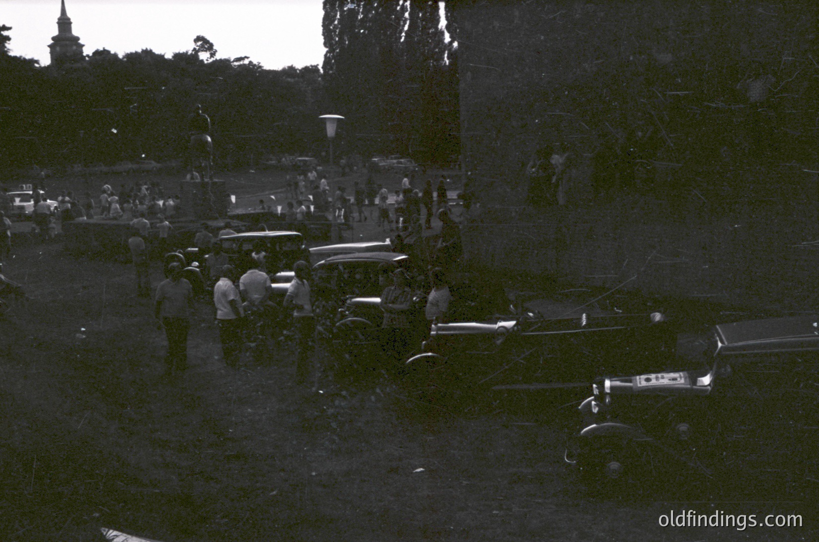 Black-and-white street scene featuring vintage cars (1940s–1950s) and a crowd gathered around a central event. Classic American automobiles, including a hearse and convertibles, parked on a tree-lined road. Low-angle lighting suggests early evening or dawn. Urban/suburban setting with visible lampposts and distant church steeple.