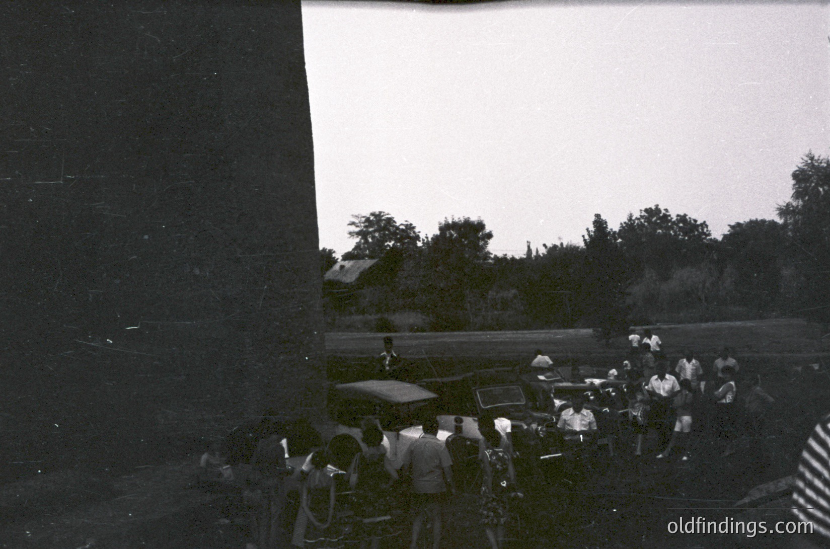 Vintage black-and-white photo of a mid-20th century outdoor gathering, likely a wedding or community event. Group of men in suits and ties seated at a long table under a tent, with a woman in a white dress standing near. Trees and a road in background suggest a rural or suburban setting.