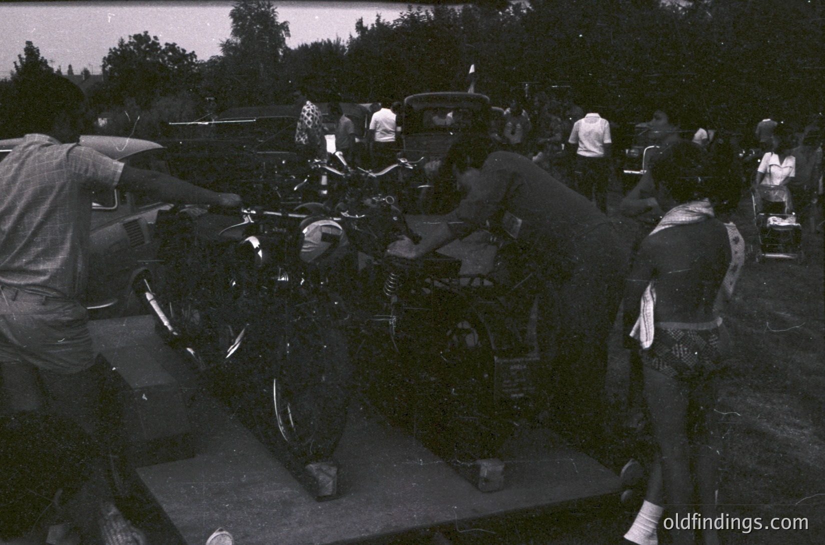 Vintage black-and-white photo of a motorcycle stunt act on a raised platform, featuring a rider in full leathers performing a wheelie. Crowd in background suggests outdoor event, likely 1960s–1970s. Classic motorcycle design and attire highlight era’s biker culture.