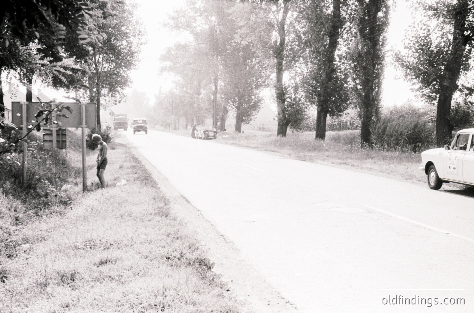 Mid-20th century rural road scene with light dusting of snow. A lone figure in winter attire leans against a fence beside a narrow, unpaved road lined with mature trees. Classic cars, including a white sedan, drive along the road, suggesting limited traffic. Snow-covered grass and minimal infrastructure hint at a quiet, possibly Eastern European countryside.