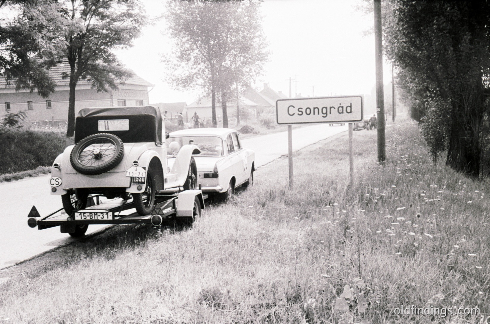 Mid-20th century roadside scene in Csongrád, Hungary. Classic vintage car (1940s-50s) towing a trailer with a newer model vehicle. Rural road flanked by grassy verge and trees. Signpost marking "Csongrád" in Hungarian script.