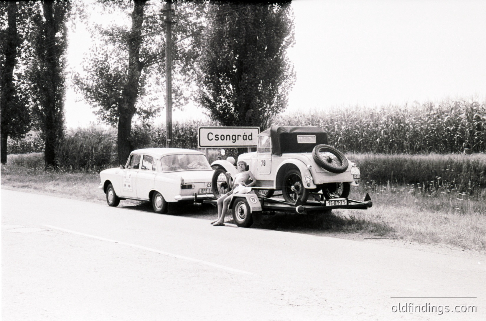 Vintage roadside scene featuring a 1950s-era car towing a classic vehicle with a flat tire. Signpost reads "Csongrád," Hungary. Rural countryside with trees and open fields in background.