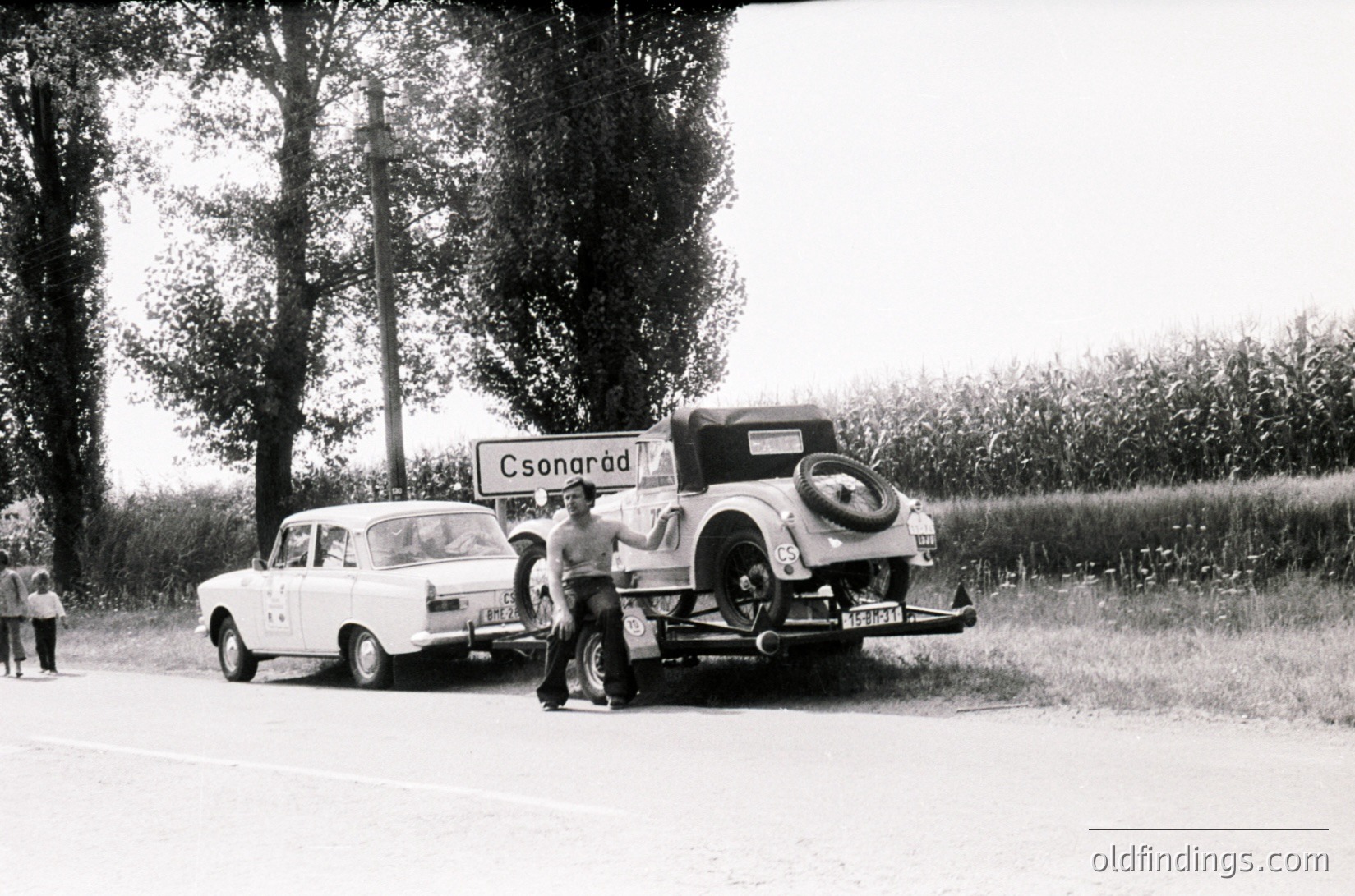 Vintage roadside scene featuring a 1960s-era white sedan towing an older classic car with a flat tire. Signpost reads "Csorarád," indicating a Hungarian location. Two men inspect the rear of the classic vehicle, while a third stands nearby. Rural road flanked by trees and open fields.