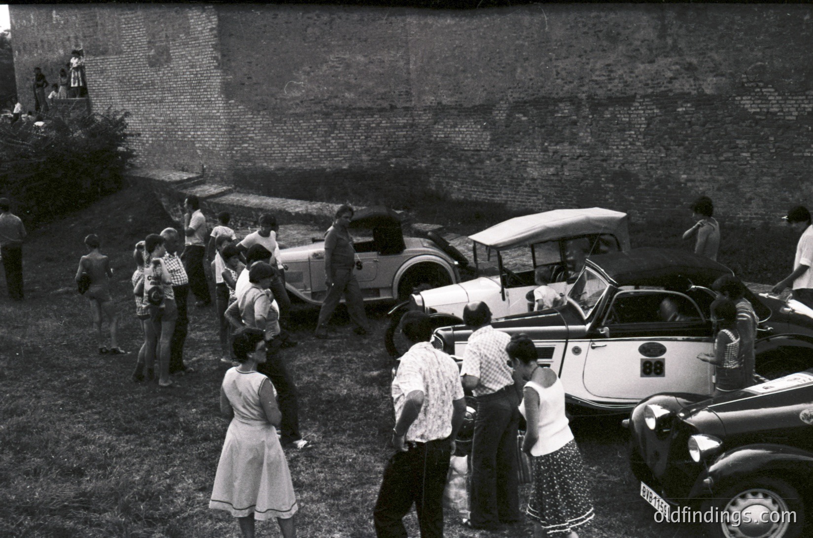 Vintage black-and-white scene featuring mid-20th-century European street life. A group of casually dressed people—men in short-sleeve shirts and women in knee-length dresses—gather near vintage convertibles and a classic car with a white top. Brick wall and greenery in background suggest an urban park or courtyard. Likely 1950s–1960s era.