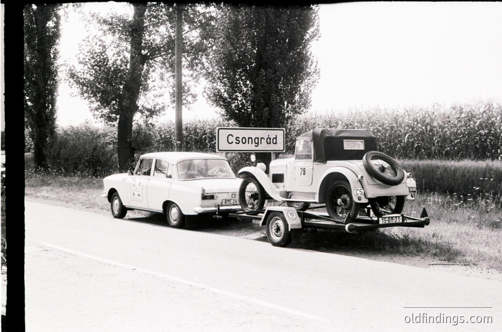 Vintage flatbed tow truck hauling a classic roadster, labeled "Csongrád" sign in background. Mid-20th century Hungarian countryside road scene.